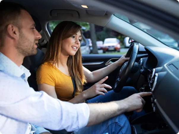 Un moniteur de conduite pointe vers le tableau de bord, la conductrice sourit.