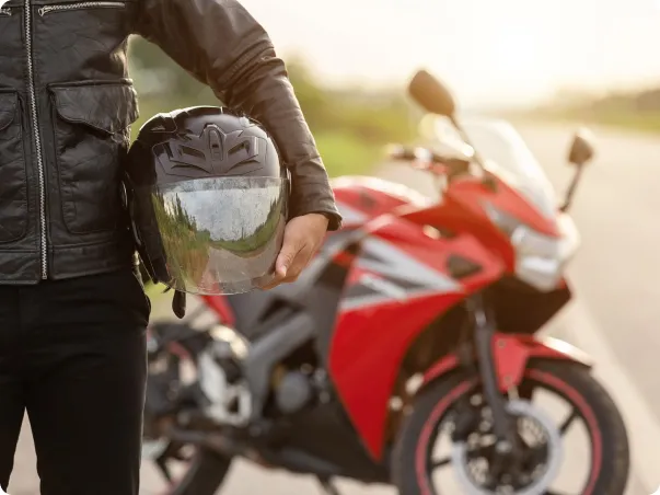 Motard en veste de cuir noir tenant un casque à côté d'une moto rouge.