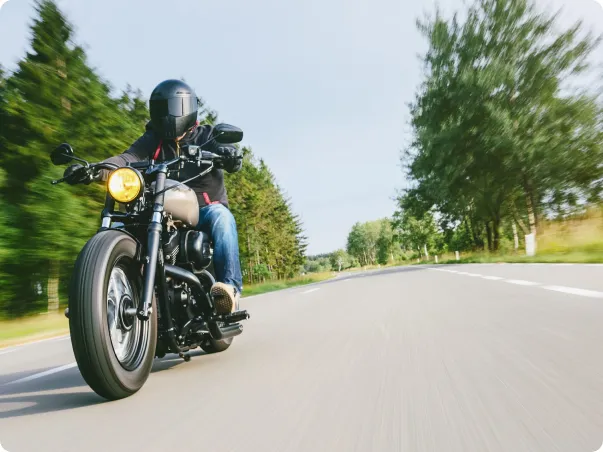 Motard casqué sur une moto noire vintage, roulant sur une route de campagne bordée d'arbres.