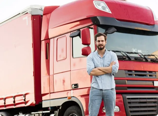 Chauffeur routier barbu debout devant un camion rouge avec les bras croisés.